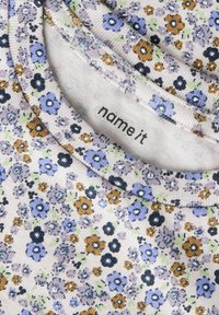 Close-up van stof met bloemenprint in paars, zwart en bruine tinten, met een "name it" merklabel in de ronde halslijn.