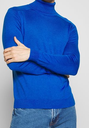 Person iført en lyseblå højhalset sweater og lyseblå jeans, med armene krydset og den ene hånd, der griber om den modsatte arm.