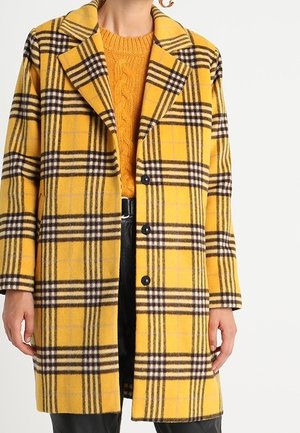 Manteau à carreaux jaunes avec grands revers porté sur un pull orange en maille torsadée, porté avec un pantalon noir et une ceinture noire.