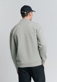 Grijze sweatshirt met een hoge kraag en ribbelboorden. Draag het met een zwarte broek en een zwarte pet met een metalen verstelknop.