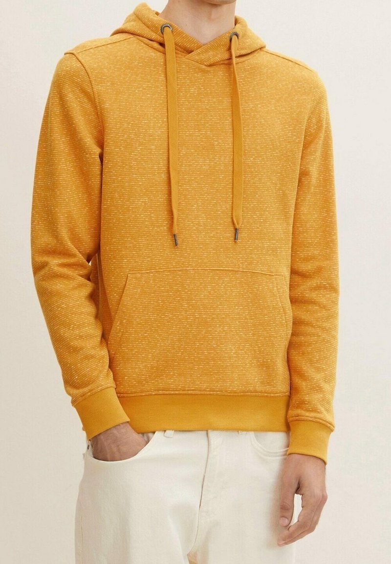 Homme portant un sweat à capuche jaune moutarde avec cordons et poche avant, associé à un pantalon blanc, sur un fond uni.