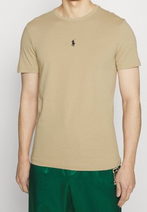 Man draagt een beige T-shirt met korte mouwen met een klein zwart logo op de borst en groene shorts met een patch aan de zijkant.