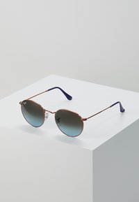 Occhiali da sole con lenti circolari a gradiente e sottile montatura in metallo color oro rosa. Terminali delle astine in plastica blu. Posizionati su un piedistallo bianco.