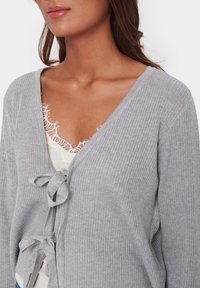 Grå ribbad cardigan med V-urringning, knytframslutning och långa ärmar, lager på lager över en krämfärgad topp med spetsdetaljer.