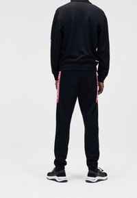 Chándal deportivo negro que incluye una chaqueta con cremallera y pantalones a juego con rayas laterales rojas en contraste. Fabricado en un tejido suave y ligero.