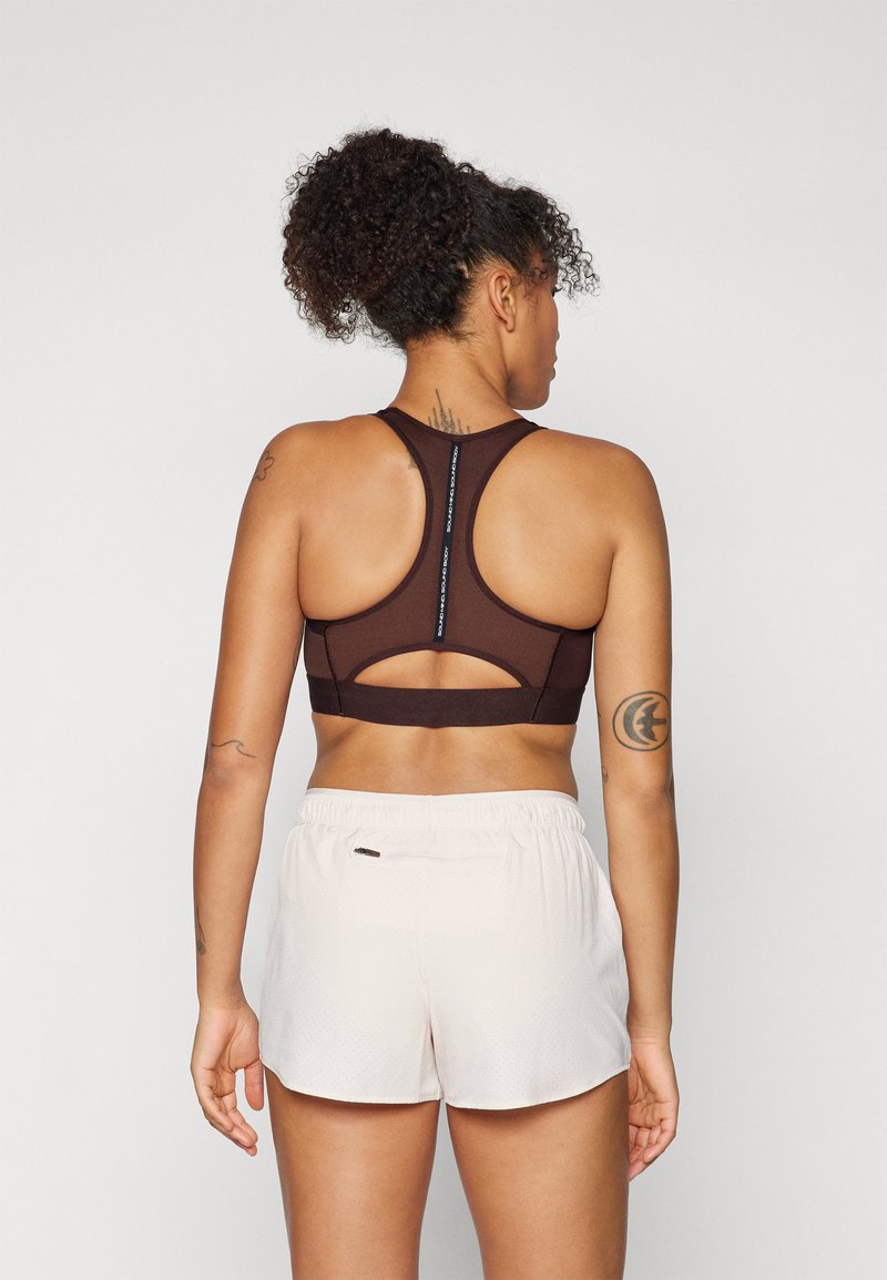 Braun-sport-BH mit Racerback-Design und einem Cutout-Detail. Kombiniert mit hellen Shorts, die ein strukturiertes Muster aufweisen.