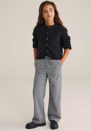 Cardigan nero a maglia con bottoni, indossato sopra pantaloni a gamba larga con fantasia pied de poule. Il modello indossa scarpe nere spesse e posa con le mani in tasca.