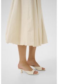 Mule beige con tacco, open toe, in pelle liscia e con un tacco a blocco basso e angolato. Indossati con una gonna fluida di colore chiaro.