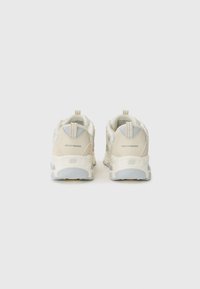 Beige sneakers with a thick white sole, featuring a textured fabric upper and a logo on the heel. Pull tabs at the back for easy wear.