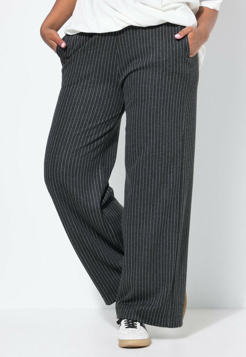 Pantalons larges en tissu gris foncé avec des rayures blanches verticales. Dotés de poches latérales et d'une coupe décontractée. Associés à des baskets blanches.