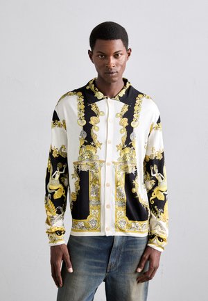 Man wearing black, white, and gold patterned button-up shirt with sea motifs and blue jeans, standing against a plain light gray background.