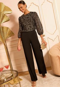 Pantaloni neri a gamba larga abbinati a un top trasparente ricamato nero con motivi a spirale dorati. Gli accessori includono una pochette dorata e sandali.