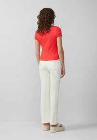 Vrouw met krullend haar staat met haar rug naar de kijker, gekleed in een strakke rode t-shirt met korte mouwen, witte broeken en beige platform sandalen.