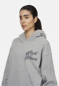Felpa grigia con cappuccio e una grande tasca frontale, con la scritta "Good for Nothing" in grigio scuro. Tessuto morbido, vestibilità comoda e cuciture resistenti.
