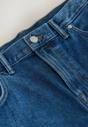 Nærbillede af taljen på blå denimjeans med sølvknap, bæltestropper og synlige syninger på lys baggrund.