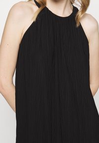 Mustat hihattomat mekko, jossa on rypistynyt rakenne, väljä leikkaus ja korkea kaula-aukko. Siinä on ohuet hihnat, ja se ulottuu olkapäiden alapuolelle.