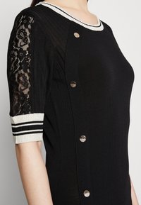 Robe en maille noire avec manches courtes en dentelle, poignets et encolure à rayures blanches et noires, et boutons décoratifs dorés le long du côté.