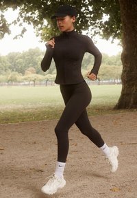 Donna che corre su un sentiero di ghiaia in un parco, indossando abbigliamento sportivo completamente nero, calzini bianchi Nike, scarpe da ginnastica bianche e un cappellino nero.