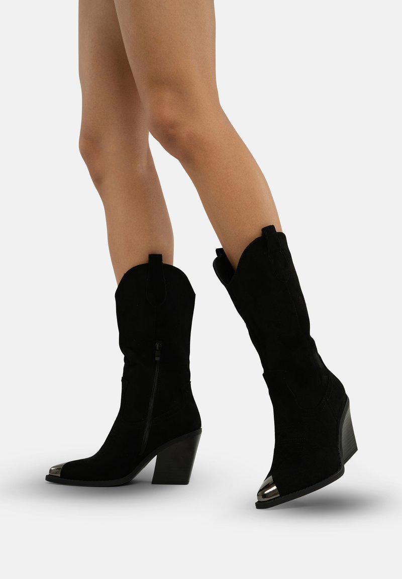Botas de vaquero de ante negro con un eje curvado, puntera metálica, cremallera lateral y tacón cuadrado. Textura suave, diseño elevado.