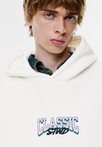 Witte hoodie met een groot meerkleurig "CLASSIC" logo in roze, groen en zwart, gedragen over een blauw denim overhemd met knoopsluiting.