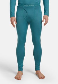 Uomo vestito con pantaloni intimi lunghi aderenti color teal con cintura con logo, in piedi a piedi nudi su uno sfondo bianco semplice.