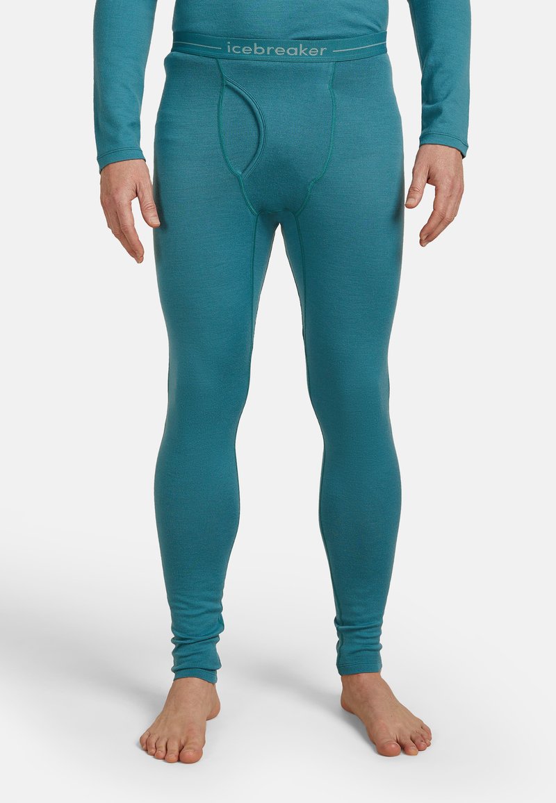Uomo vestito con pantaloni intimi lunghi aderenti color teal con cintura con logo, in piedi a piedi nudi su uno sfondo bianco semplice.