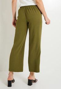 Pantalons larges vert olive en tissu texturé, avec une taille élastique et des poches latérales, associés à des sandales à talons blocs noires.