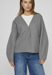Grå, avslappnad stickad tröja i mjukt material med V-ringning, sänkta axlar och långa ärmar. Stylad över en vit topp.