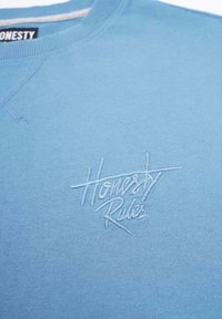 Ljusblå sweatshirt i bomull, med ribbad halsringning och manschetter. Broderad vit text meddelar "Ärlighet Regler" längst ner till höger.