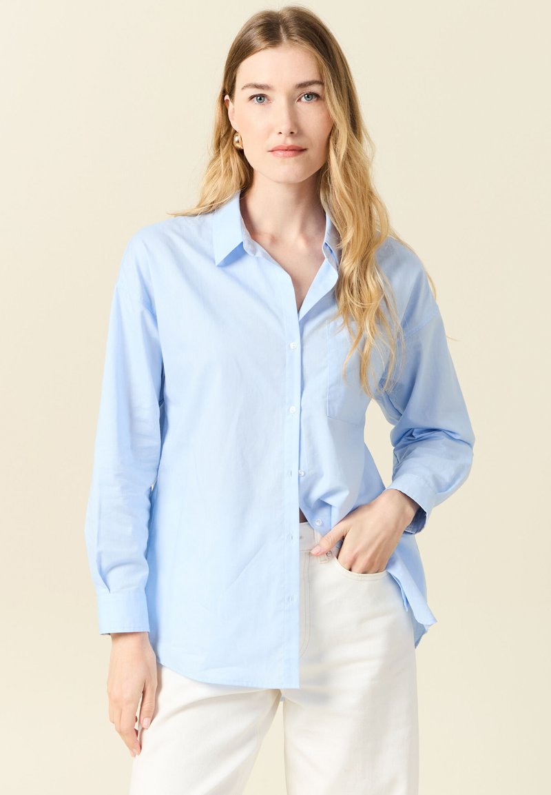 Camicia azzurro chiaro con maniche lunghe, taschino sul lato sinistro del petto e orlo dritto. Realizzata in un tessuto liscio e tessuto.