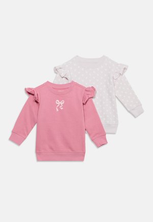 Deux chemises à manches longues pour enfants avec des épaules volantées, une rose avec un motif de nœud, une blanche avec un motif de cœur.