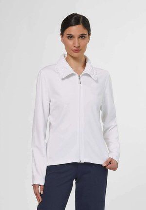 Femme portant une veste blanche zippée avec un col texturé et un pantalon bleu marine, debout devant un fond clair uni.