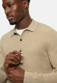 Beige Strukturstrick-Polohemd mit Kragen, schwarzen Knöpfen und langen Ärmeln. Die Hände sind vor dem Hemd zusammengelegt.