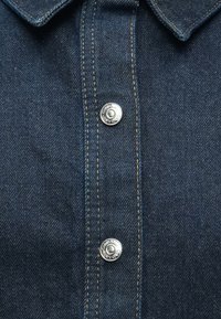 Σκούρο μπλε πουκάμισο από τζιν με μυτερό γιακά, ασημένια μεταλλικά κουμπώματα με πατ που κλείνουν, και πορτοκαλί ραφές κατά μήκος του μπροστινού μέρους και των άκρων.