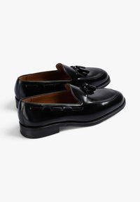 Mocasines de cuero negro con un diseño elegante, acentos de borlas, punteras redondeadas y un tacón bajo apilado. Textura suave con costuras detalladas.