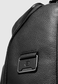 Mochila de cuero negro con superficie texturizada, con un accesorio de metal plateado brillante y una cremallera visible.