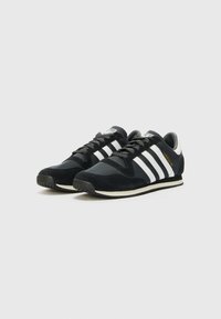 Chaussures de sport noires avec trois bandes blanches, en matériau en suède et en mesh, bout arrondi et semelle en caoutchouc texturé. Accent logo doré sur le côté.