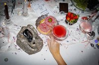 Szklane świeczniki z palącymi się białymi świecami, torebka zdobiona cekinami, kolorowe pączki z posypką, dwa koktajle, świeże truskawki i rozsypane gwiazdki.