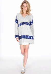 Lichtgrijze oversized trui met blauwe paillettenstrepen, V-hals en lange mouwen, gecombineerd met een zilveren rok en sneakers.