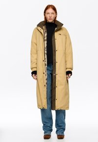 Mujer con un abrigo largo beige de invierno con ribete de piel, suéter negro, jeans azules y botas marrones, de pie contra un fondo blanco.