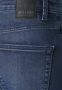 Jeans din denim albastru închis, cu o suprafață texturată, design cu cinci buzunare și o eticheta din piele neagră cu logo-ul mărcii embosat pe talie.