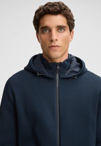 Giacca con zip di colore blu navy, caratterizzata da un motivo testurizzato, con cappuccio e fodera interna a contrasto. Visibile hardware della cerniera liscia.