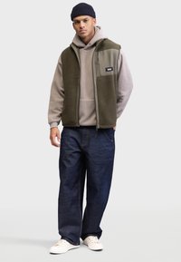 Zielona kamizelka z polaru noszona na szarym hoodie, w zestawieniu z ciemnoniebieskimi dżinsami i białymi sneakersami. Kamizelka ma kieszeń na zamek i teksturowany materiał.