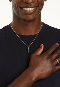 Zilveren ketting met een rechthoekige hanger met een zwarte steeninleg, liggend op een donker T-shirt, met een hand op de borst geplaatst.