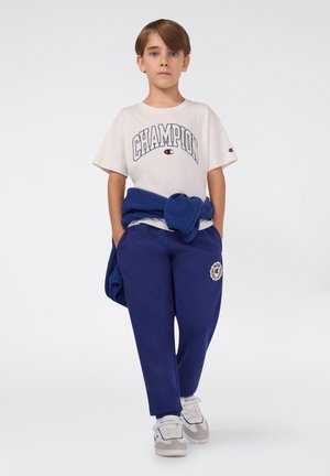 Biała koszulka z grafiką "CHAMPION", niebieskie dresy z naszywką logo, niebieski sweter przewiązany w talii, jasne sneakersy z szarymi akcentami.