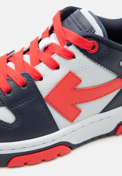 Witte en marineblauwe sneaker met felrode veters, een grote rode pijl aan de zijkant en een gevoerde rode en marineblauwe zool.