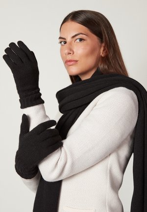 Guanti e sciarpa neri a maglia, con polsini a coste e una texture morbida, indossati con un maglione bianco a trama. Design semplice e monocromatico.