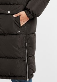 Svart pufferjacka med en ficka på sidan som stängs med dragkedja och en knappknäppning. Tyget är glatt och något glänsande, med kviltade partier.