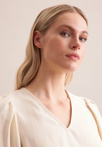Blusa crema a V con texture morbida, spalle strutturate e un delicato drappeggio. Il tessuto morbido e la naturale lucentezza aumentano l'eleganza.