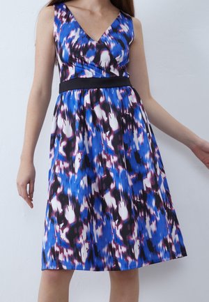 Sukienka bez rękawów do kolan w abstrakcyjny wzór w kolorach niebieskim, białym, czarnym i fioletowym, z czarnym paskiem w talii, noszona przez stojącą osobę.